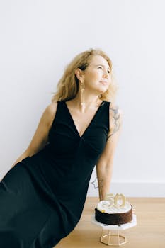 Blonde woman in black dress posing beside a 50th birthday cake indoors.