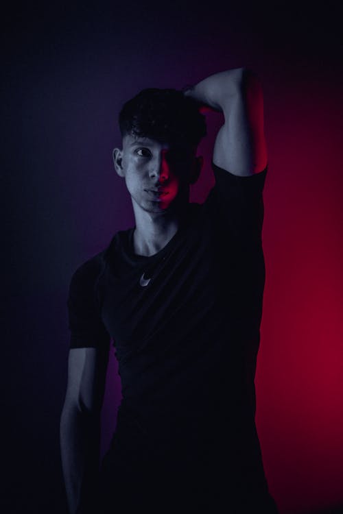 Free Confident young male in black outfit standing with raised arm and looking at camera in dark room in neon light Stock Photo