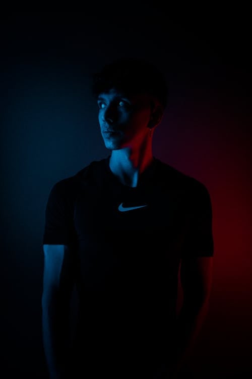 Free Pensive young male in black t shirt looking away while standing in darkness with neon light in studio Stock Photo
