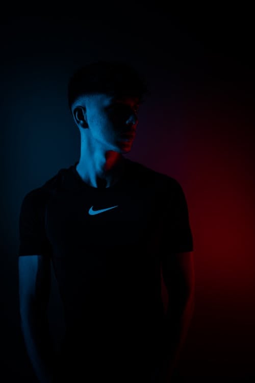 Dreamy handsome male in casual clothes looking away while standing near wall in darkness with glowing neon illumination