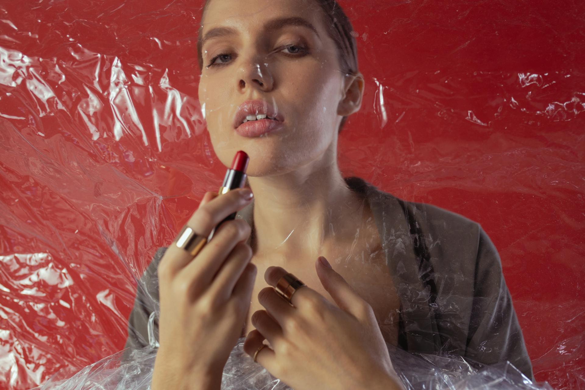 Artistic studio shot of a woman applying lipstick, wrapped in plastic, against a red backdrop.