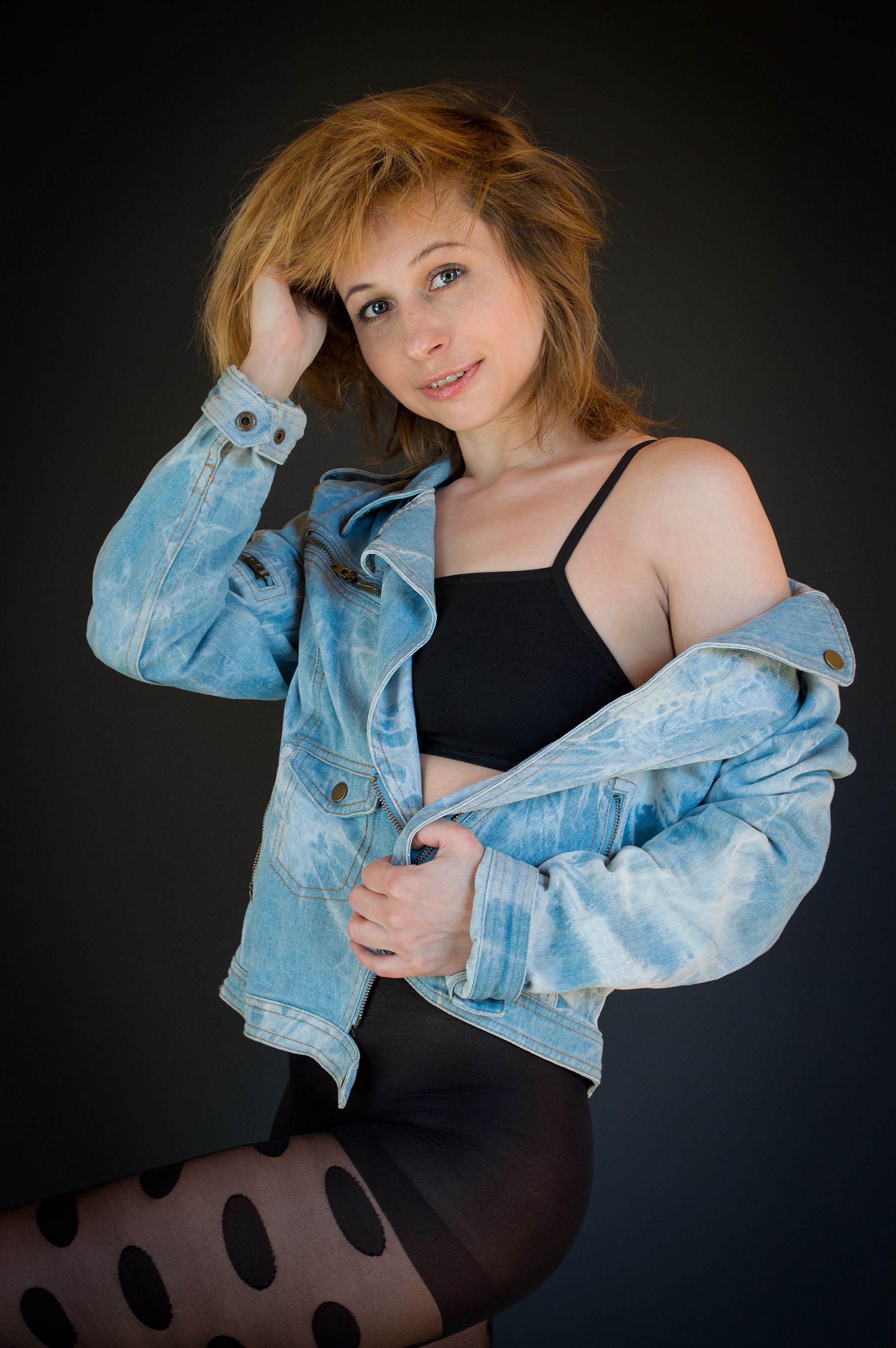 Portrait of a woman posing confidently in a denim jacket and black attire against a studio backdrop.