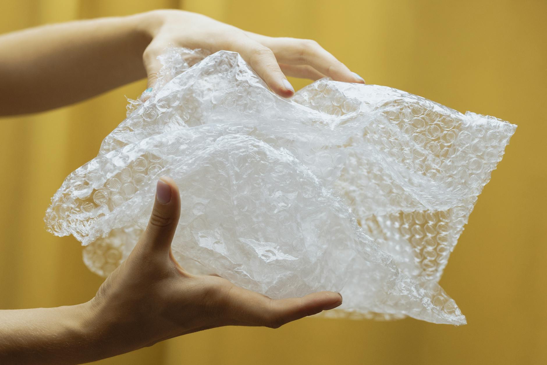 Close-up of hands holding and crumpling bubble wrap against a yellow backdrop indoors.