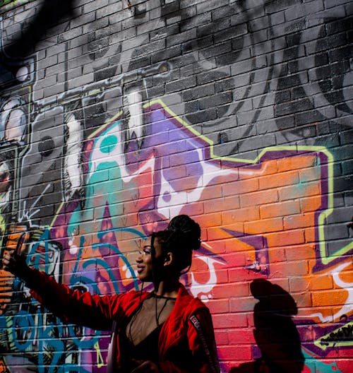 A Woman in Red Jacket Taking Selfie while Standing Near Graffiti Wall