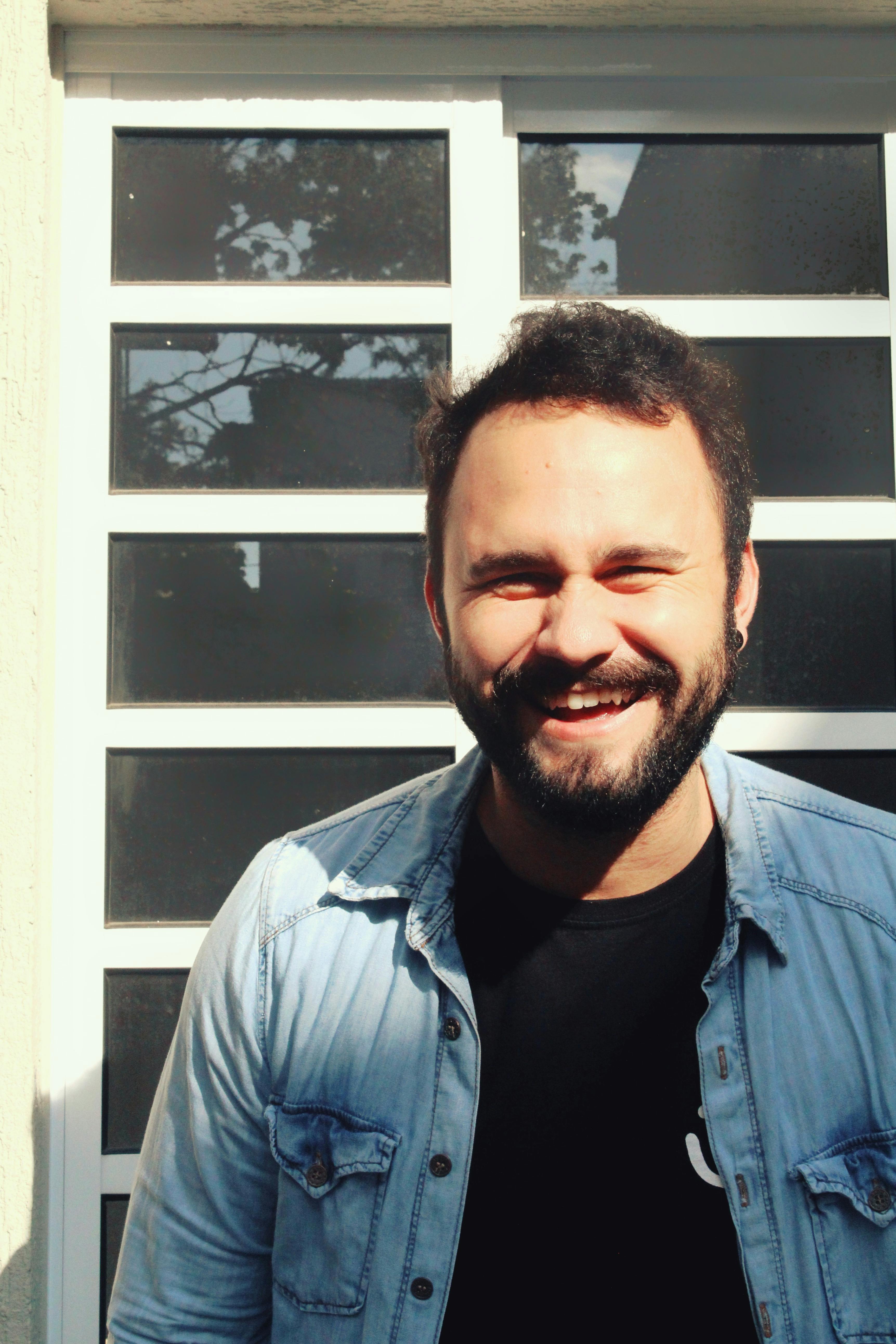 A happy man in a denim jacket and black shirt smiling brightly outdoors.