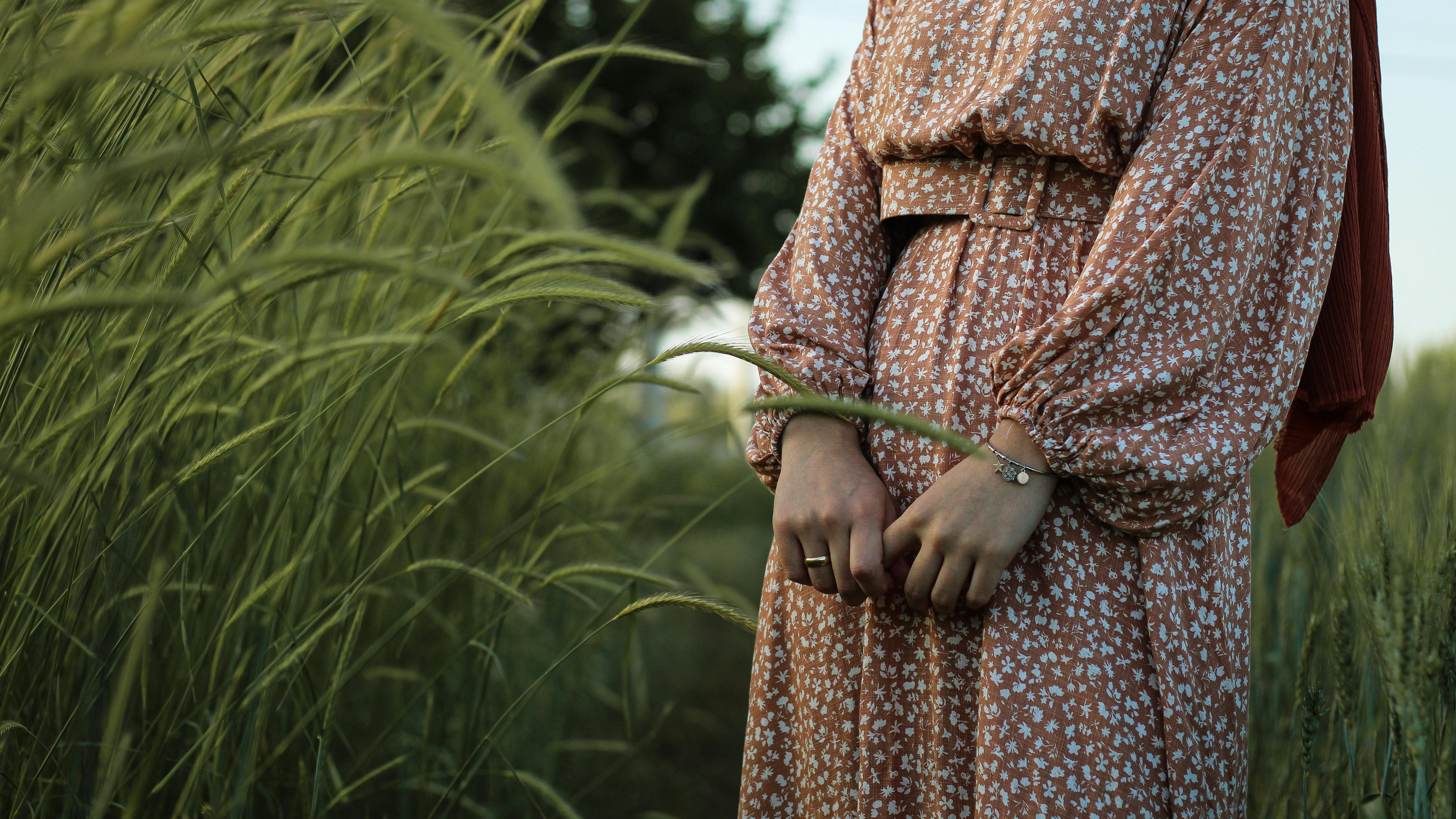 Unrecognizable crop female in boho dress standing in green meadow among tall grass in countryside
