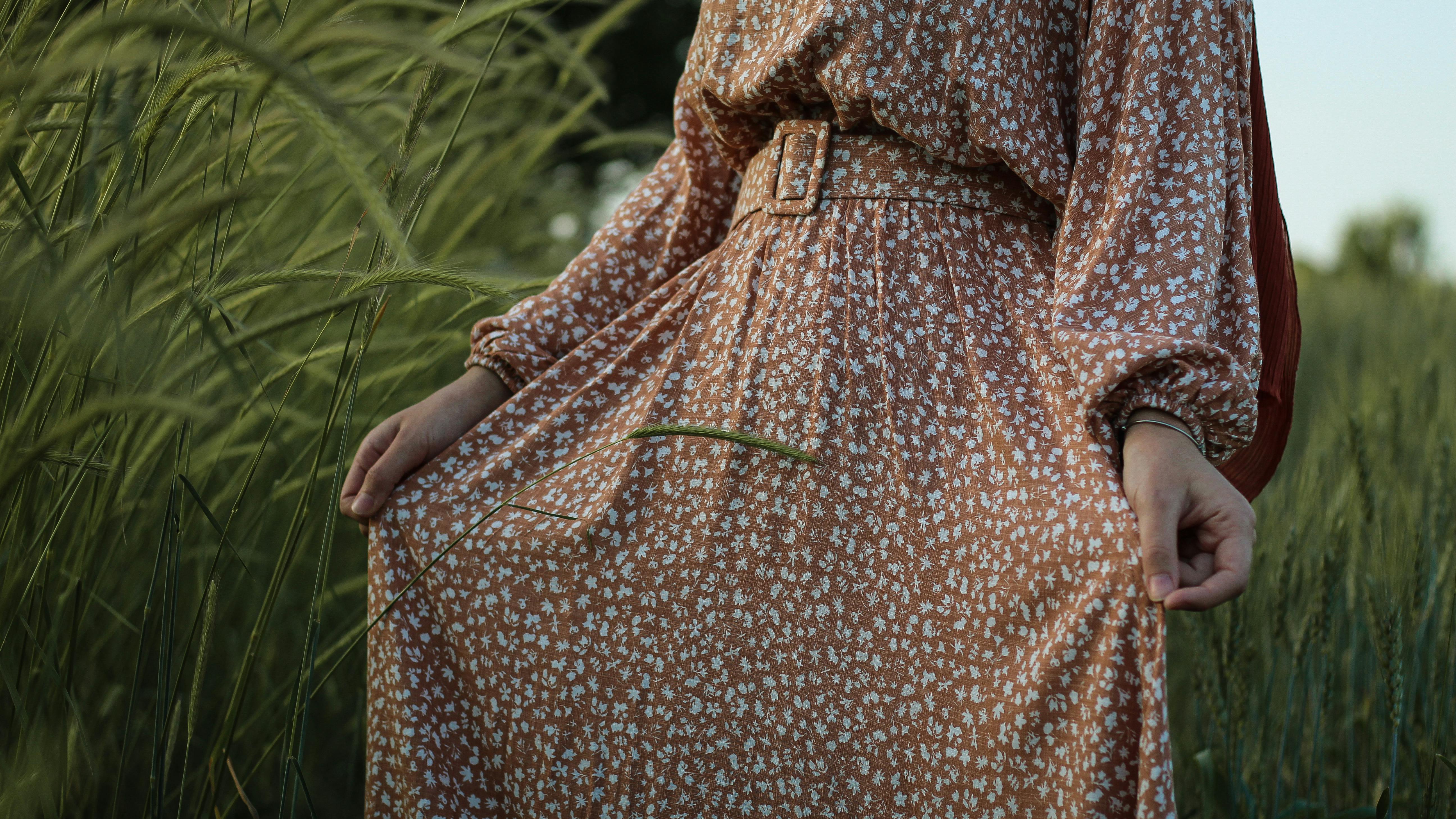 Unrecognizable crop female in spacious dress standing in green grassy field near tall grass and holding hem of dress on sunny day