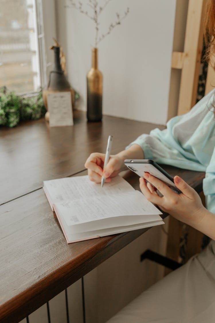 Crop Woman With Smartphone Writing In Notebook At Home