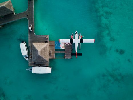 Drone view of white seaplane parked on translucent blue ocean water near small bungalow on sunny day
