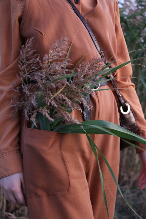 Unrecognizable crop female in orange outfit standing in field with blooming wildflowers in pocket