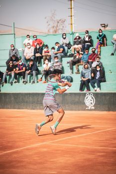 A tennis player in action on a clay court with a lively audience watching under bright daylight.