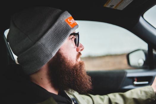 Profile of a bearded man wearing a beanie and sunglasses driving a car.