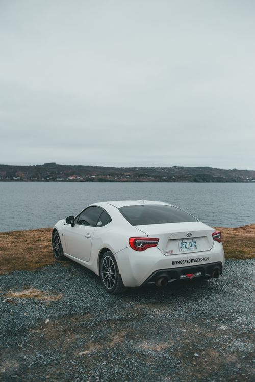 Free White luxury coupe car parked by a serene lakeside with overcast sky, showcasing elegance and tranquility. Stock Photo