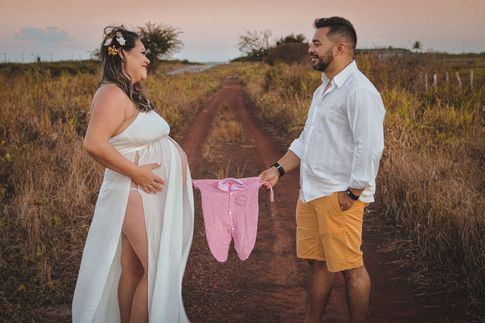 Expectant couple celebrates pregnancy in a rural setting, holding baby clothes with joy and anticipation.