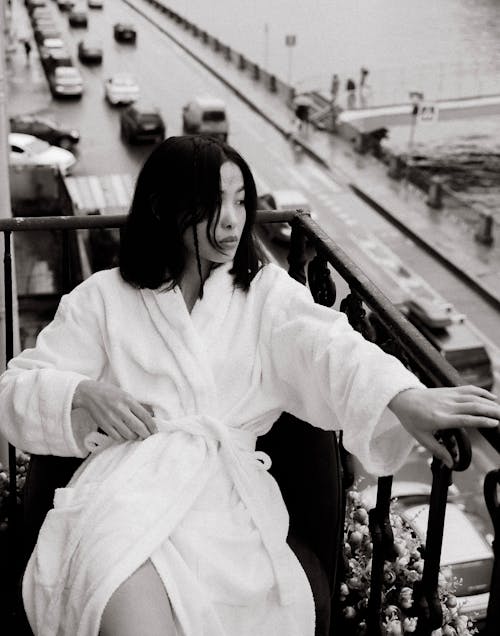 Black and white of thoughtful young female model in white bathrobe sitting on balcony above road with cars near water and looking away