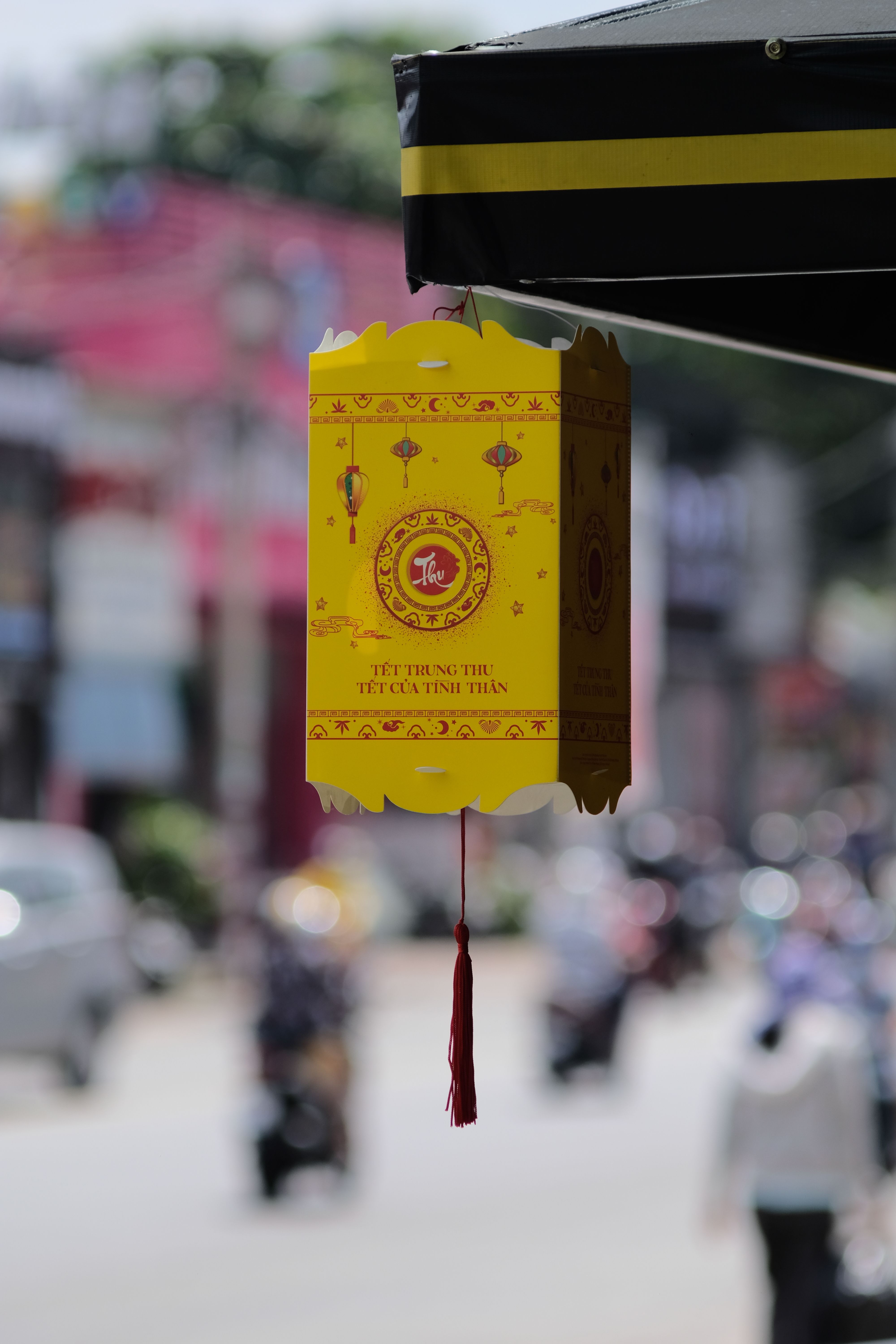 A close-up of a yellow paper lantern with traditional designs hanging outdoors, featuring a blurred urban background.