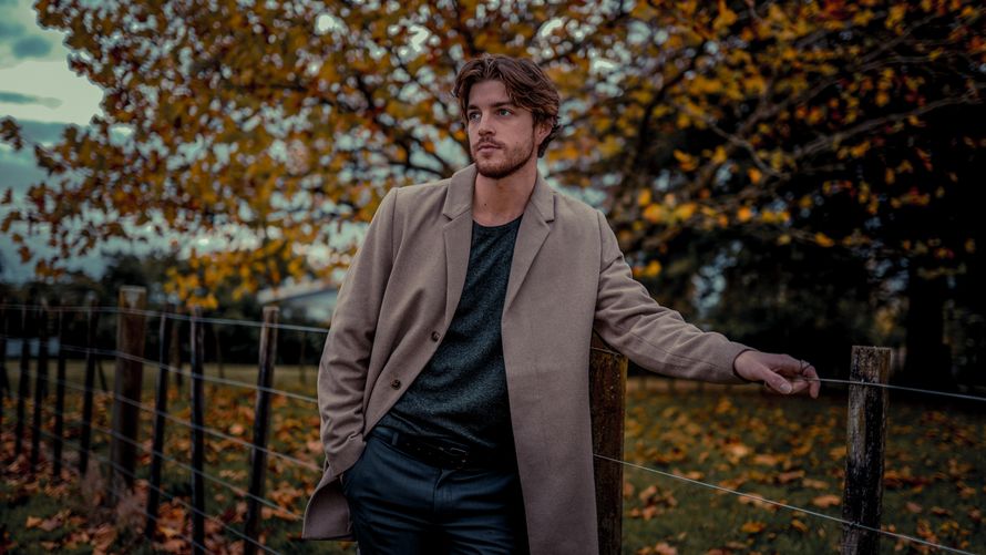 Man in a beige wool overcoat by a fence in autumn — photo by Mushtaq Hussain on Pexels