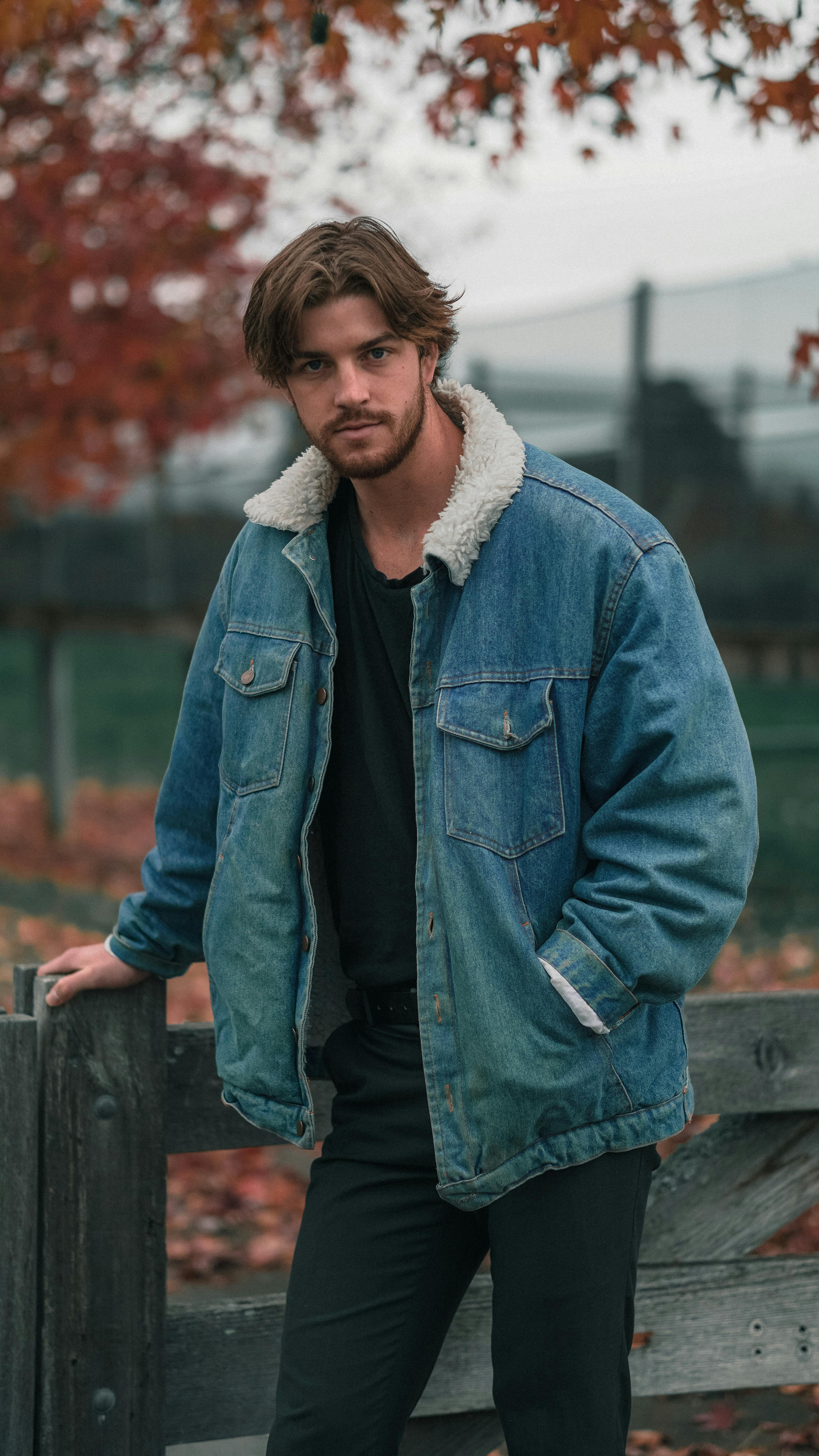 A young man wearing a denim jacket stands near a wooden fence surrounded by autumn foliage.