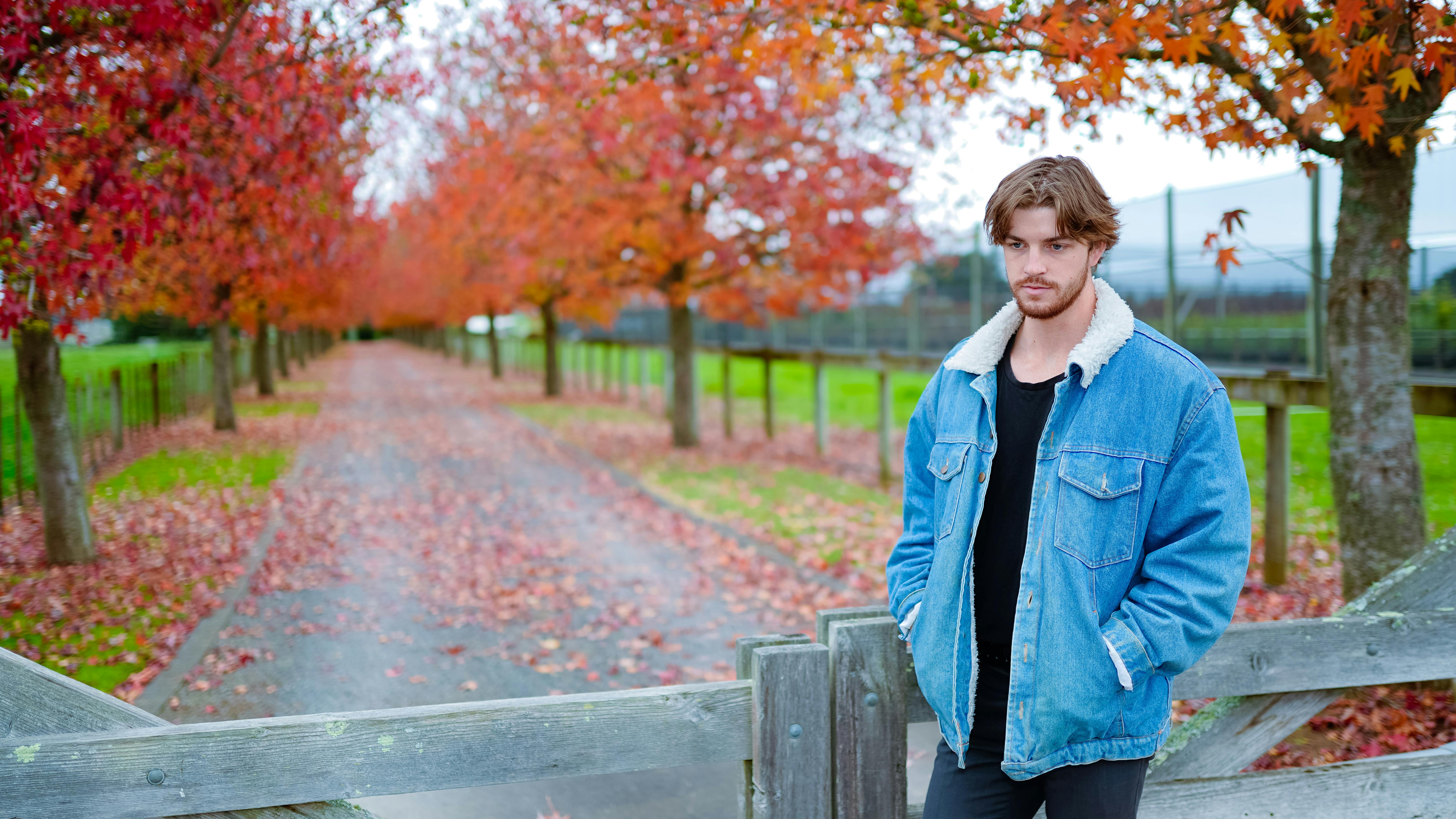Young man in a denim jacket posing outdoors amidst fall foliage.