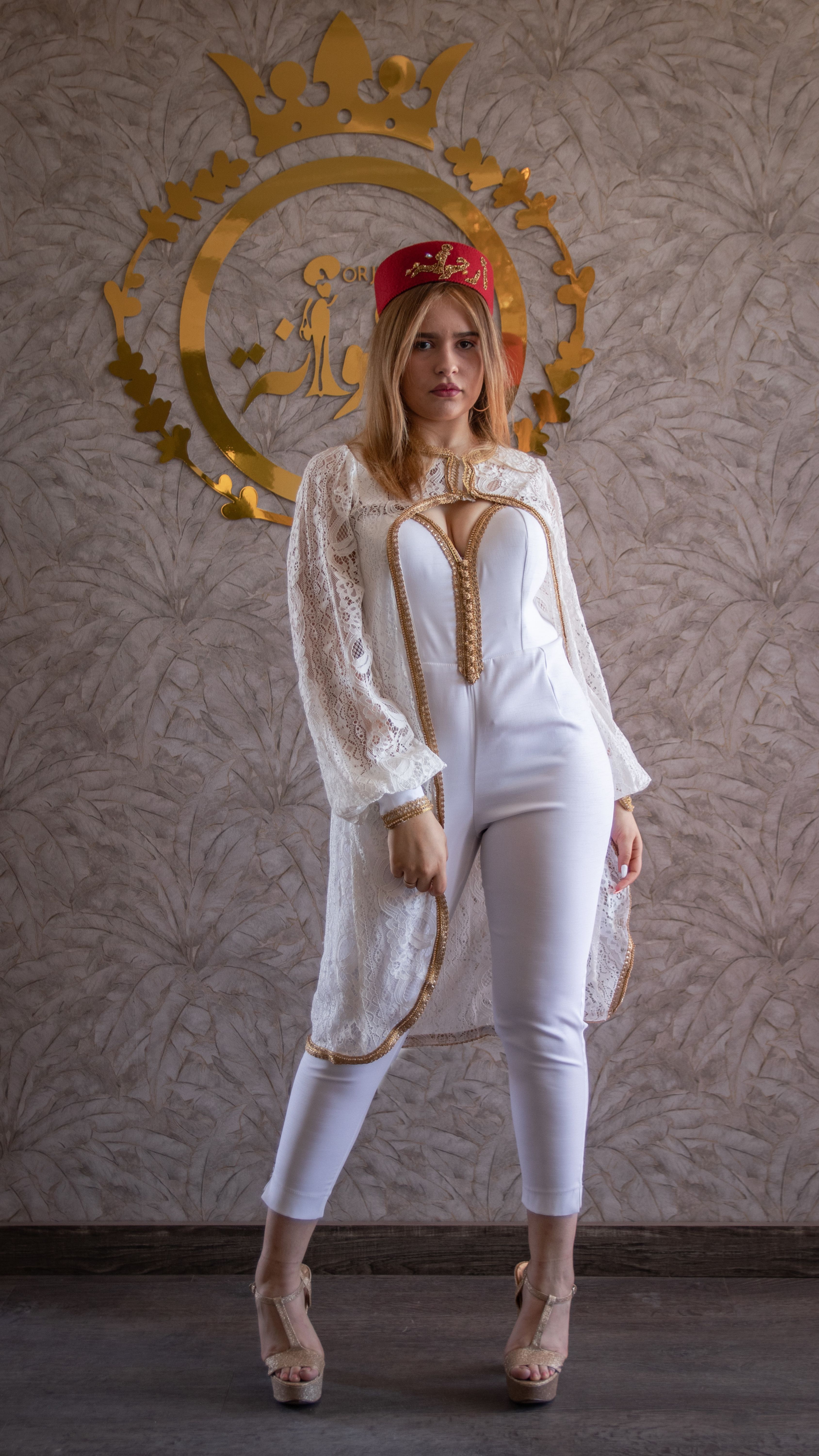 Stylish woman posing indoors in traditional cap and elegant white outfit, exuding modern fashion charm.