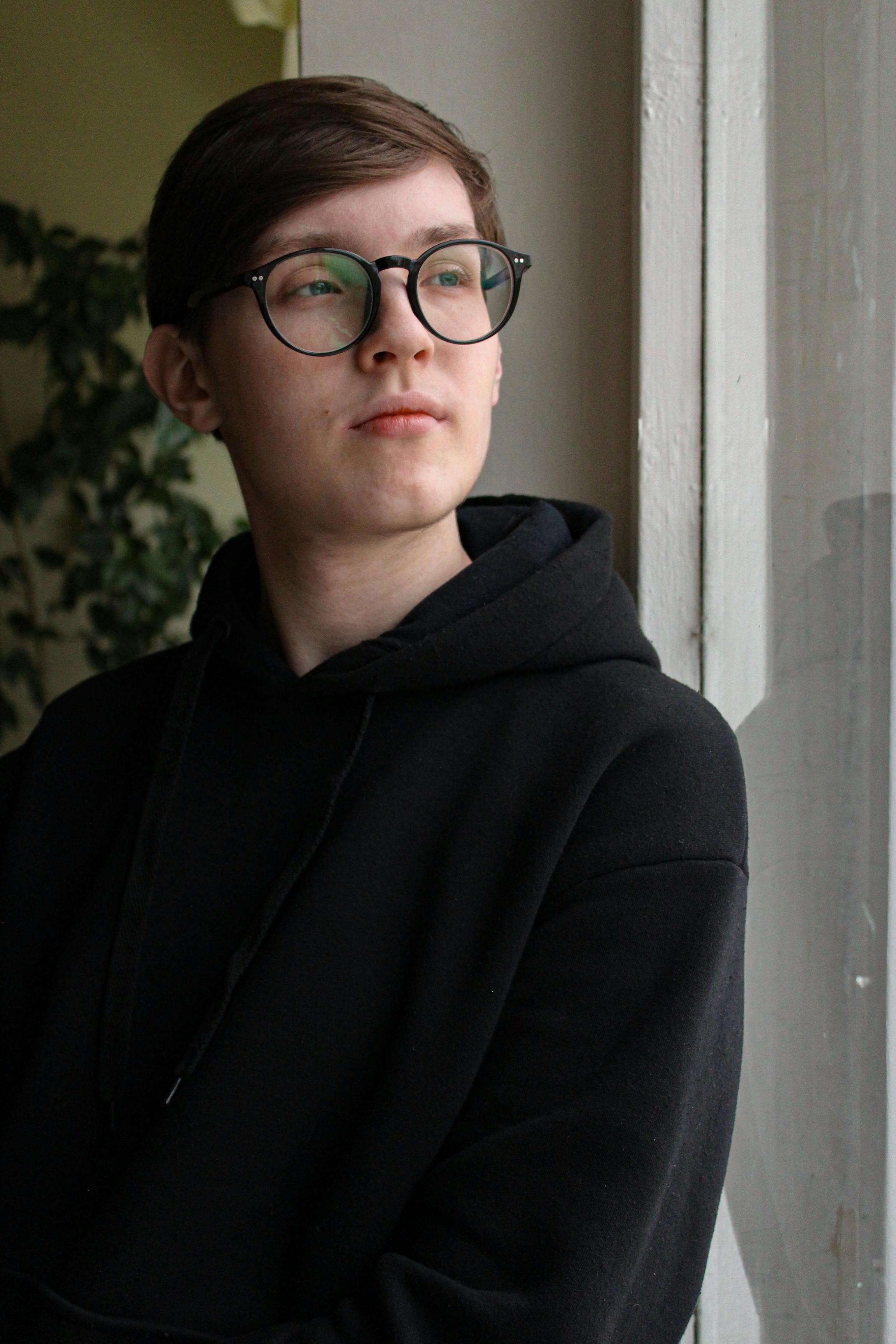 Caucasian young adult with eyeglasses wearing a hoodie, gazing thoughtfully out the window.