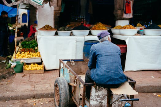 A man sits on a cart in a vibrant street market in Essaouira, Morocco, showcasing local produce and spices.