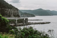 在多云的天空下，悬崖与山脉旁的沿海桥梁呈现出壮丽的景色，尽显自然之美