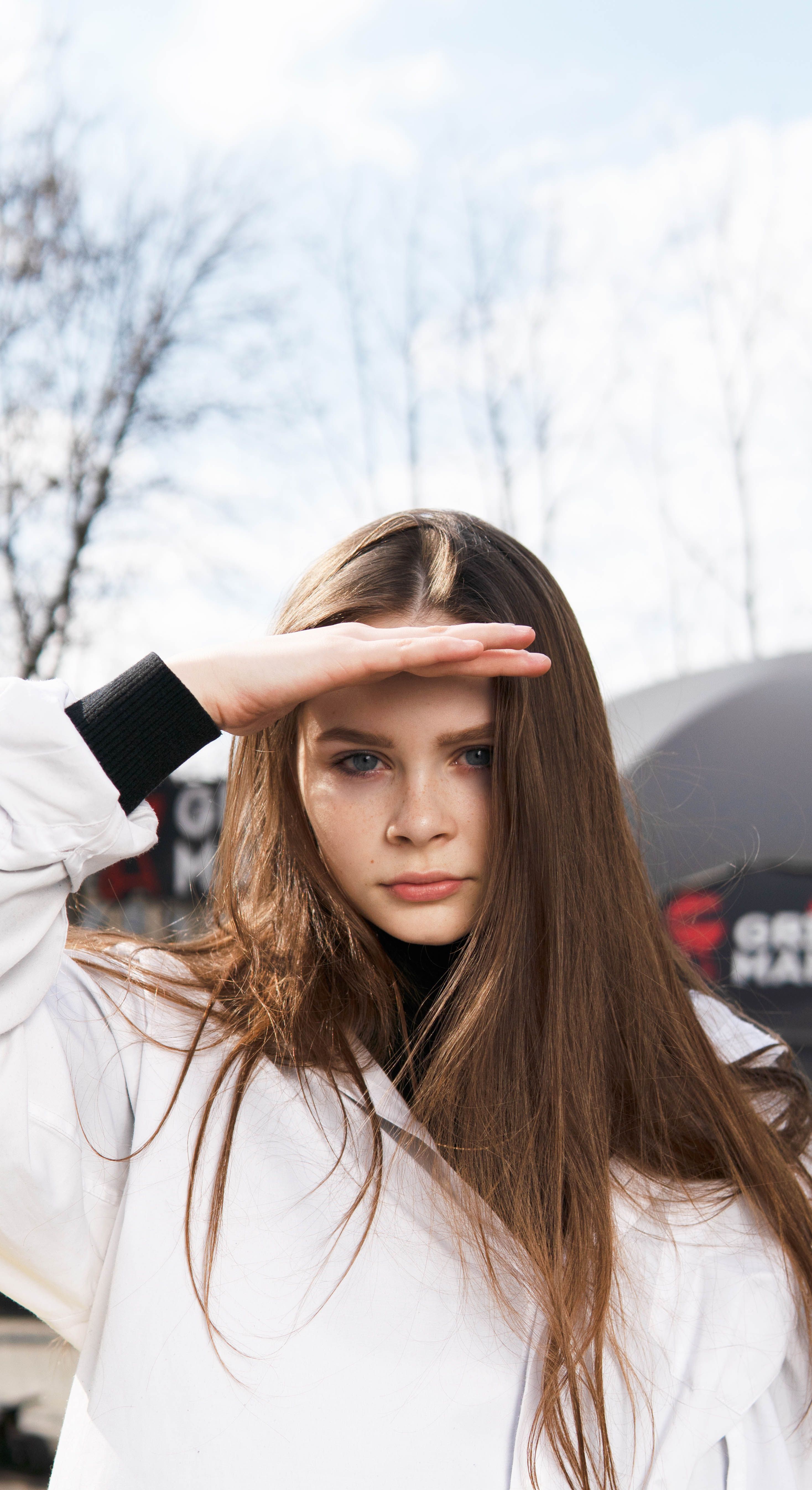 A caucasian girl with brown hair outdoors, posing with hand on forehead, in a selective focus shot.