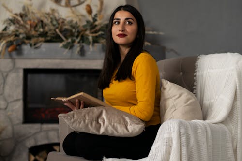A Woman in Yellow Long Sleeves Reading a Book while Sitting on the Sofa