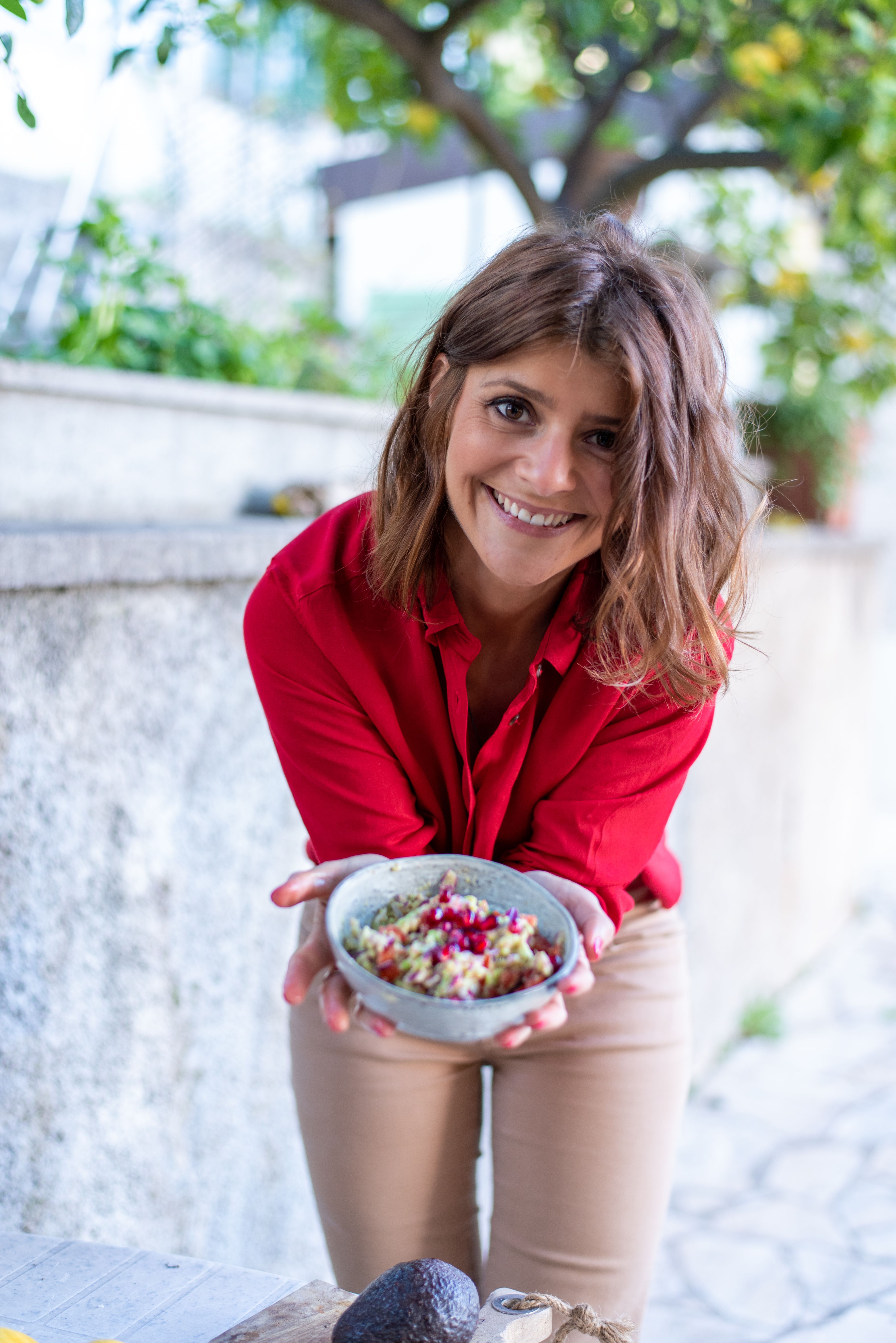 A woman in a red shirt smiling outdoors while holding a bowl of fresh salad, offering a joyful culinary experience.