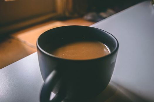 温暖的灯光下，一个黑色的杯子里盛着舒适的早晨咖啡，放在桌子上