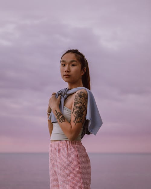 Tattooed ethnic teen against sea under cloudy sky at sunset