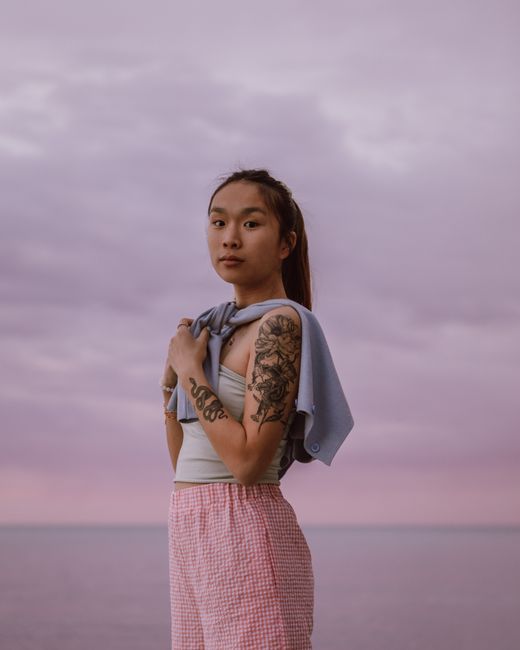 Side view of cool Asian female teenager with tattoos looking at camera against ocean under purple sky at sundown
