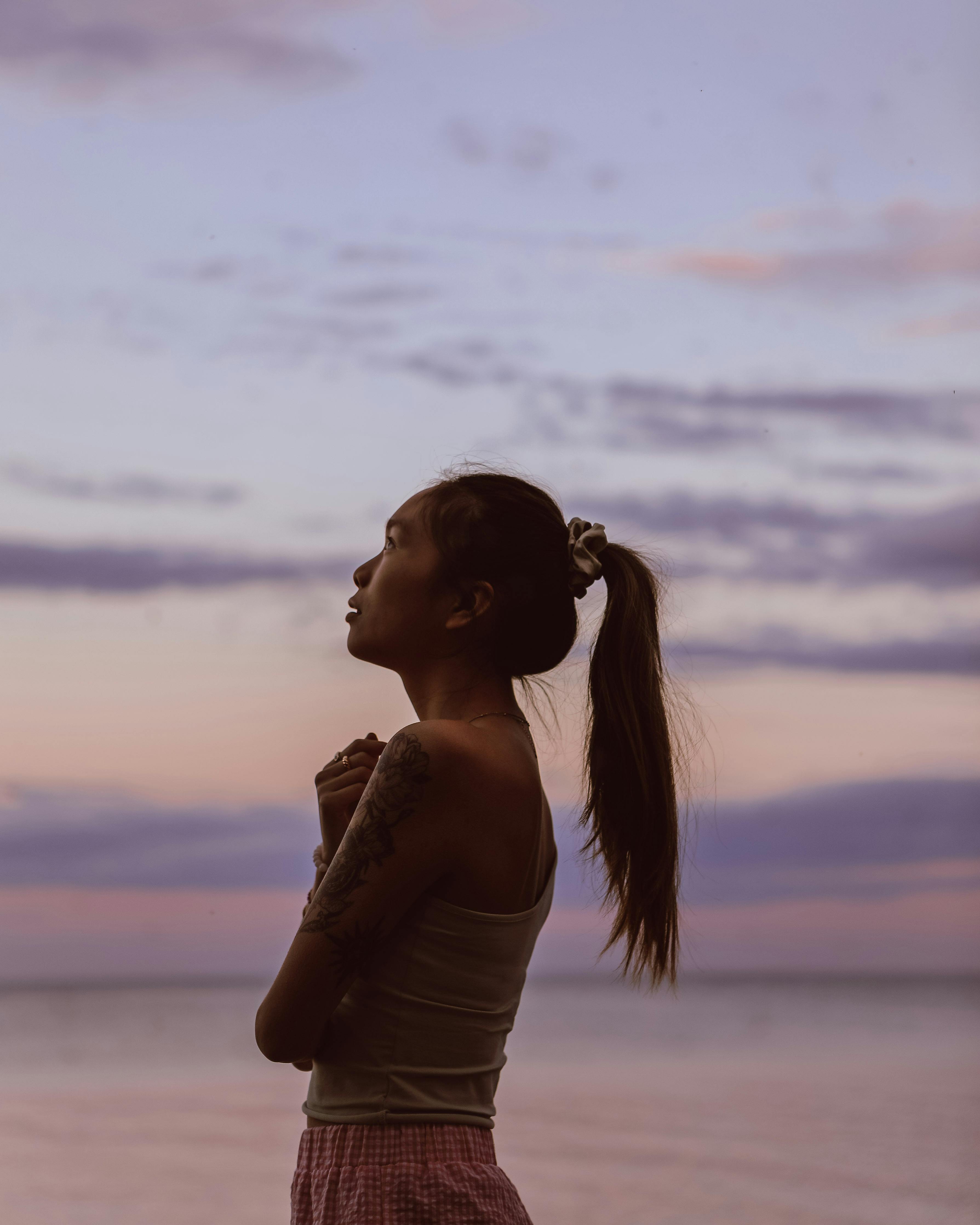 Side view of ethnic female looking up while standing against sundown sky on coast of sea in evening