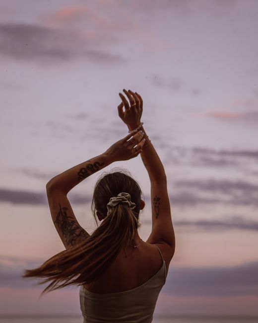 A young tattooed woman raises her arms against a serene sunset sky, creating a peaceful outdoor scene.