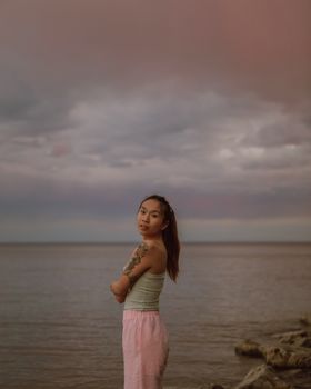 Young woman with tattoos standing by the tranquil ocean shore under a dramatic cloudy sky.
