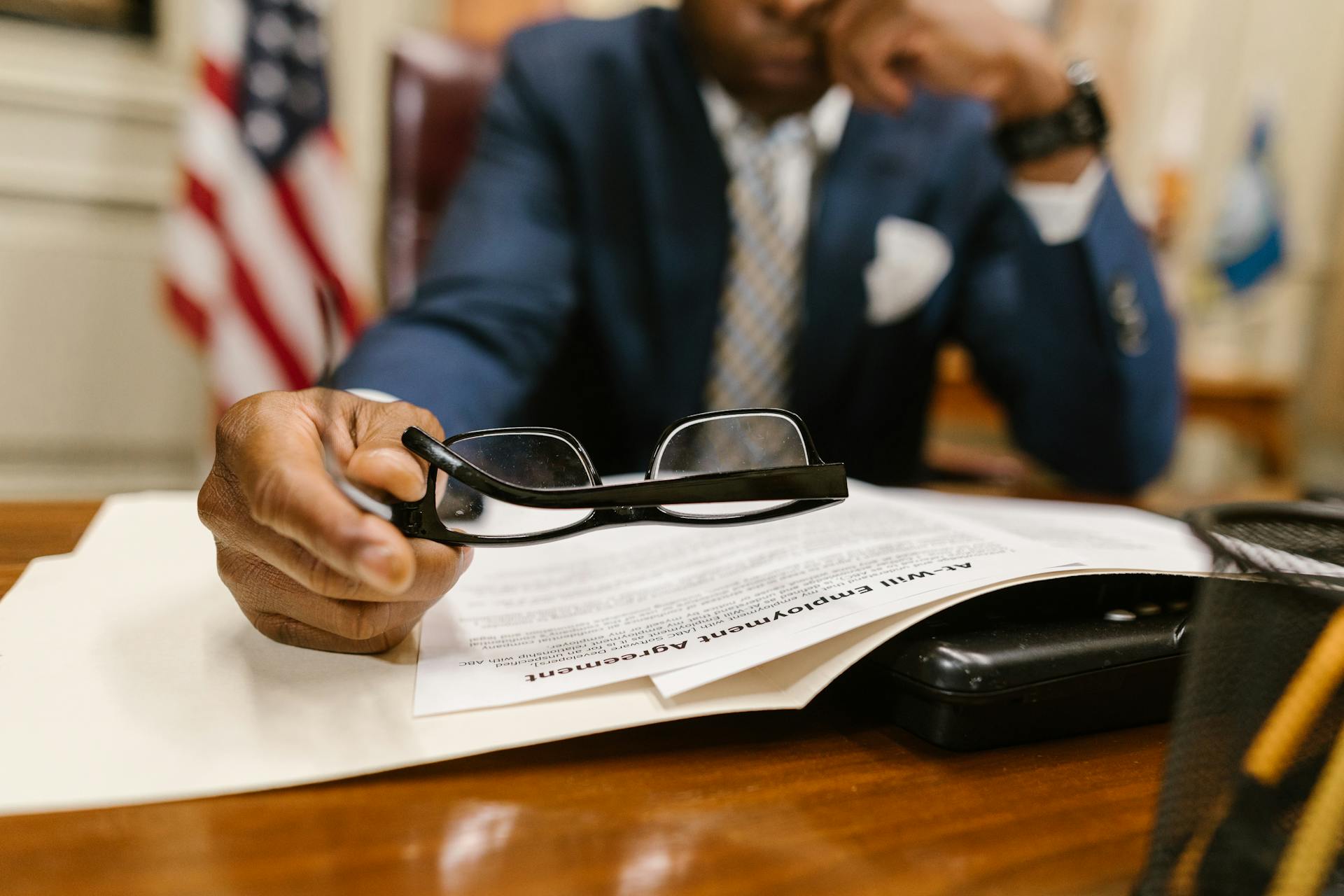 Business professional contemplating a contract, wearing a suit, seated at a desk with documents and eyeglasses.