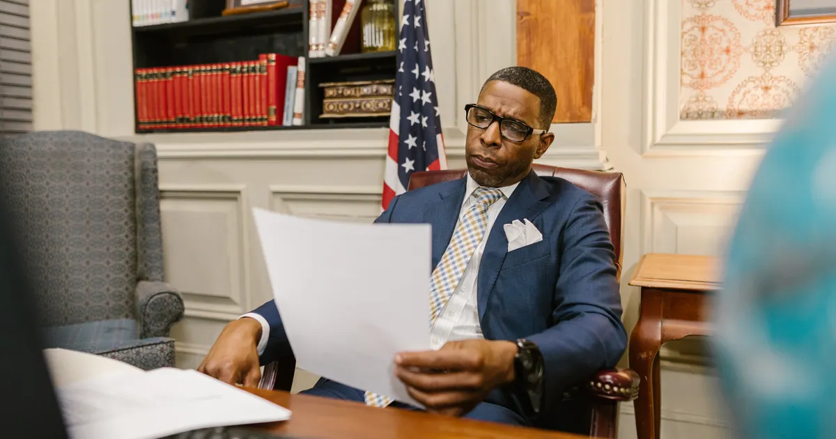 A man in a blue suit holding a document in an office with an American flag behind him, representing federal AI preemption