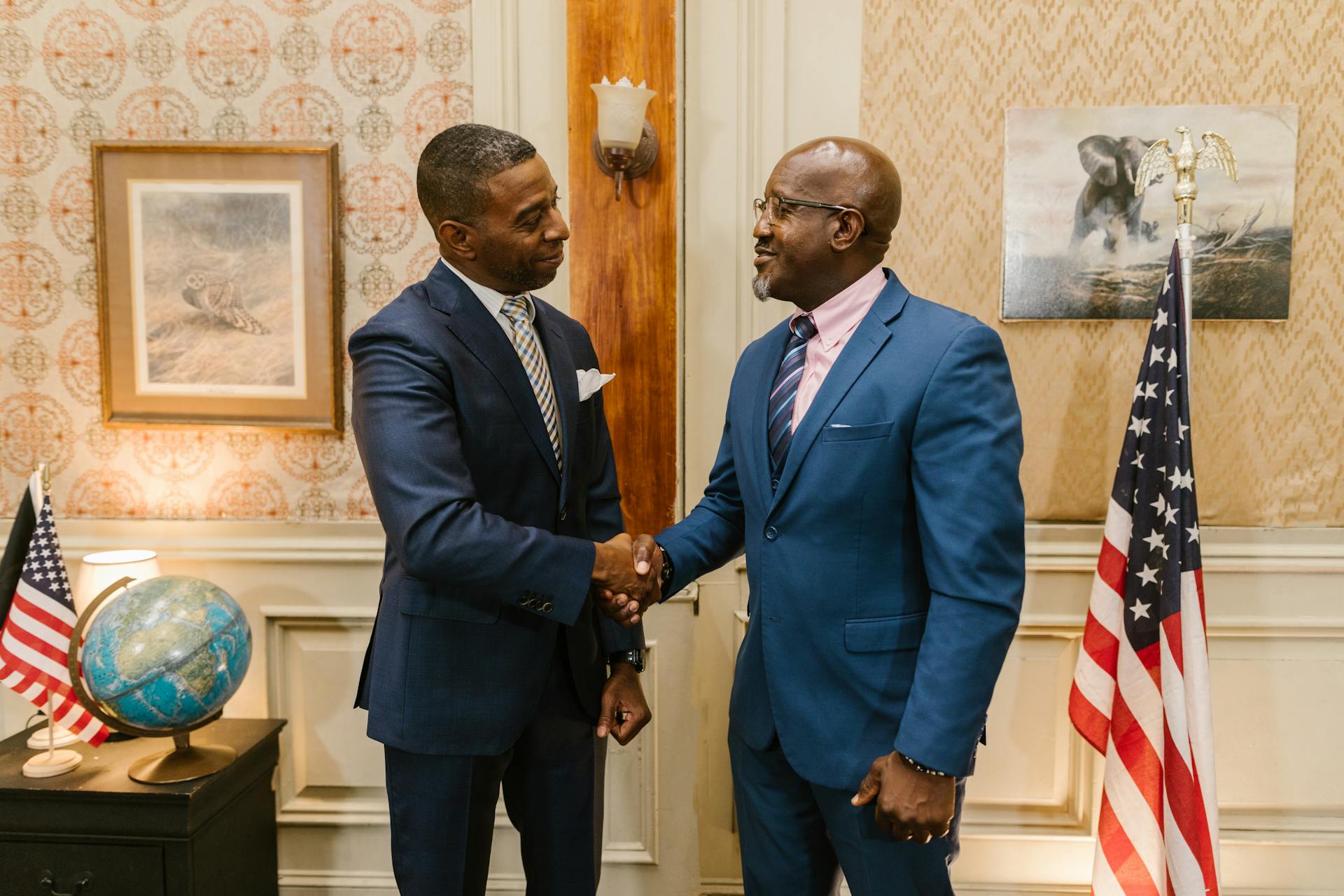 African American men in suits shaking hands in a formal law office setting with USA flags and globe.