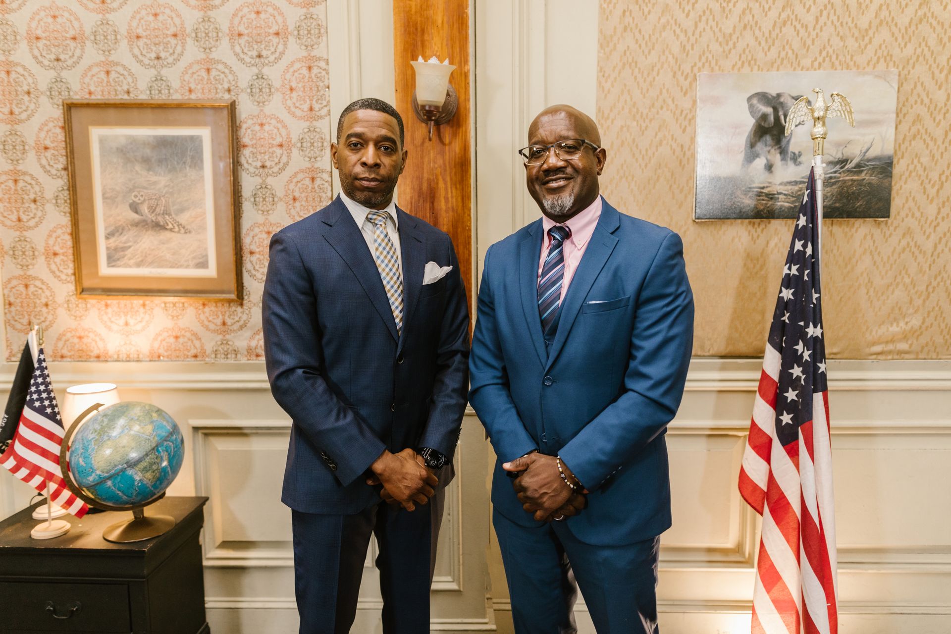 Two businessmen in suits standing in an office setting beside an American flag.