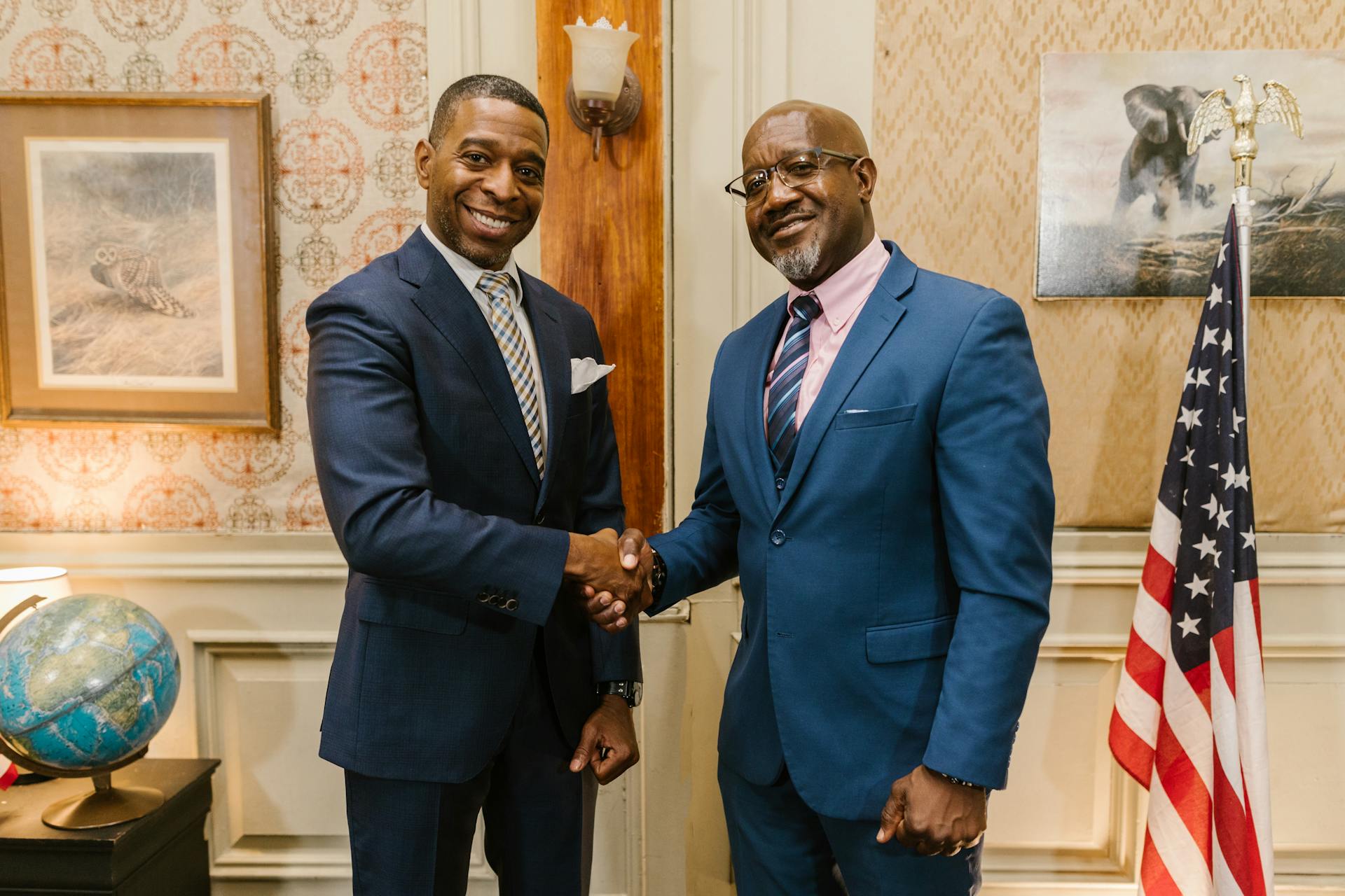 Two businessmen in suits shaking hands in an office setting with a US flag and globe.