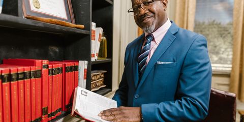 Professional lawyer in formal attire holding a book, smiling in an office setting surrounded by law books.