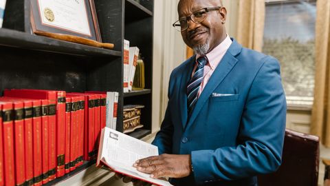 Professional lawyer in formal attire holding a book, smiling in an office setting surrounded by law books.
