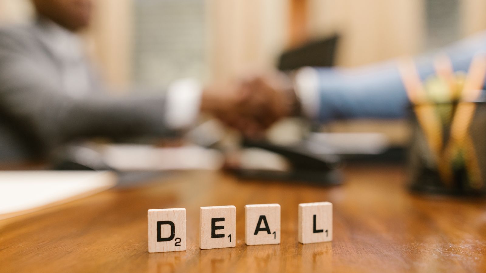 Close-up of Scrabble tiles spelling 'deal' with a handshake in the background.