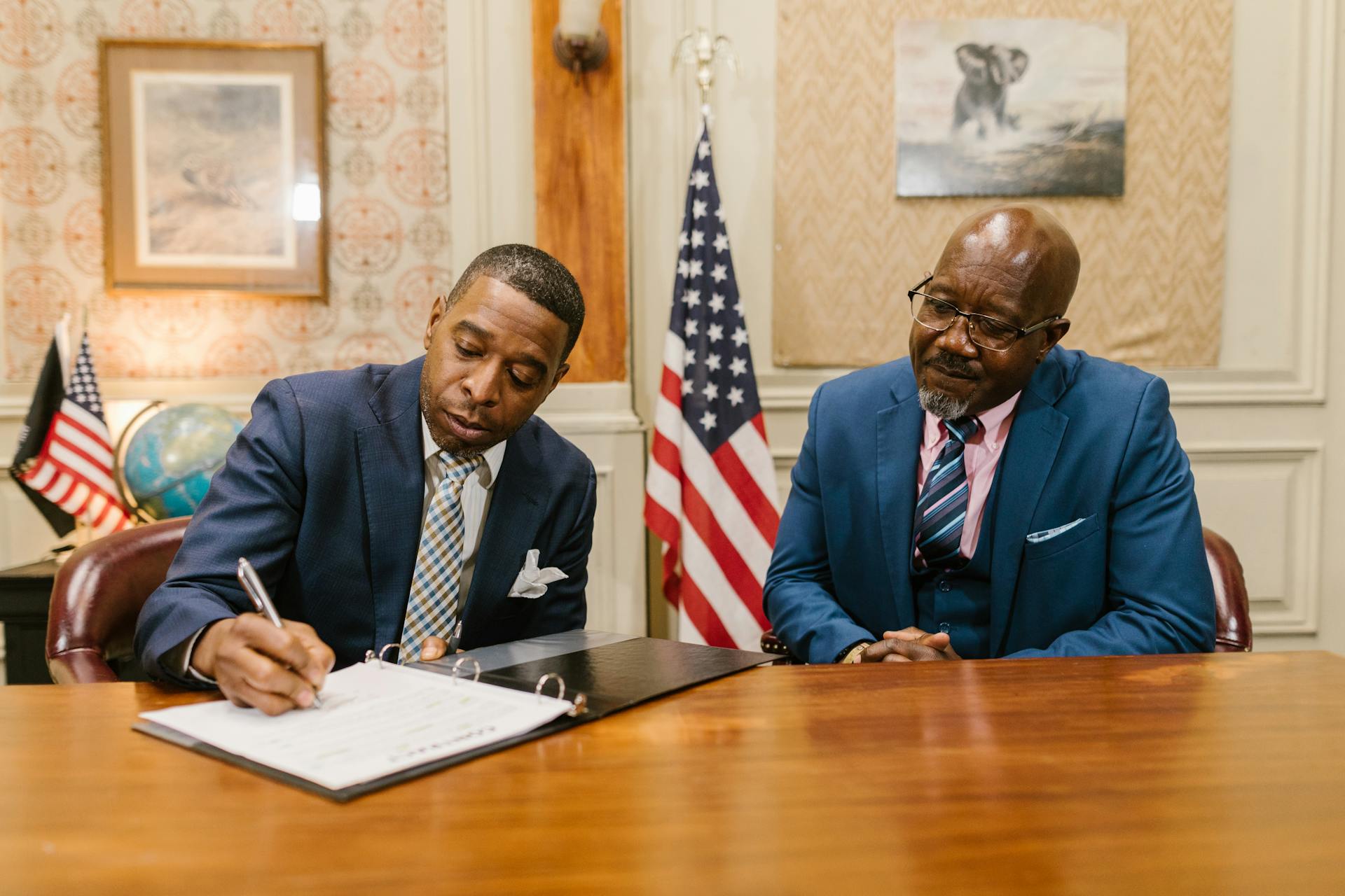 Two businessmen in suits sitting at a table signing documents in a formal office setting.