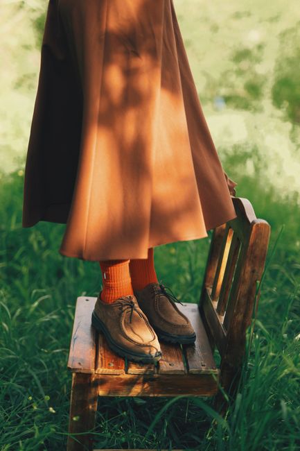 Crop unrecognizable woman in stylish skirt and shoes standing on wooden chair on green grass in nature in sunny day