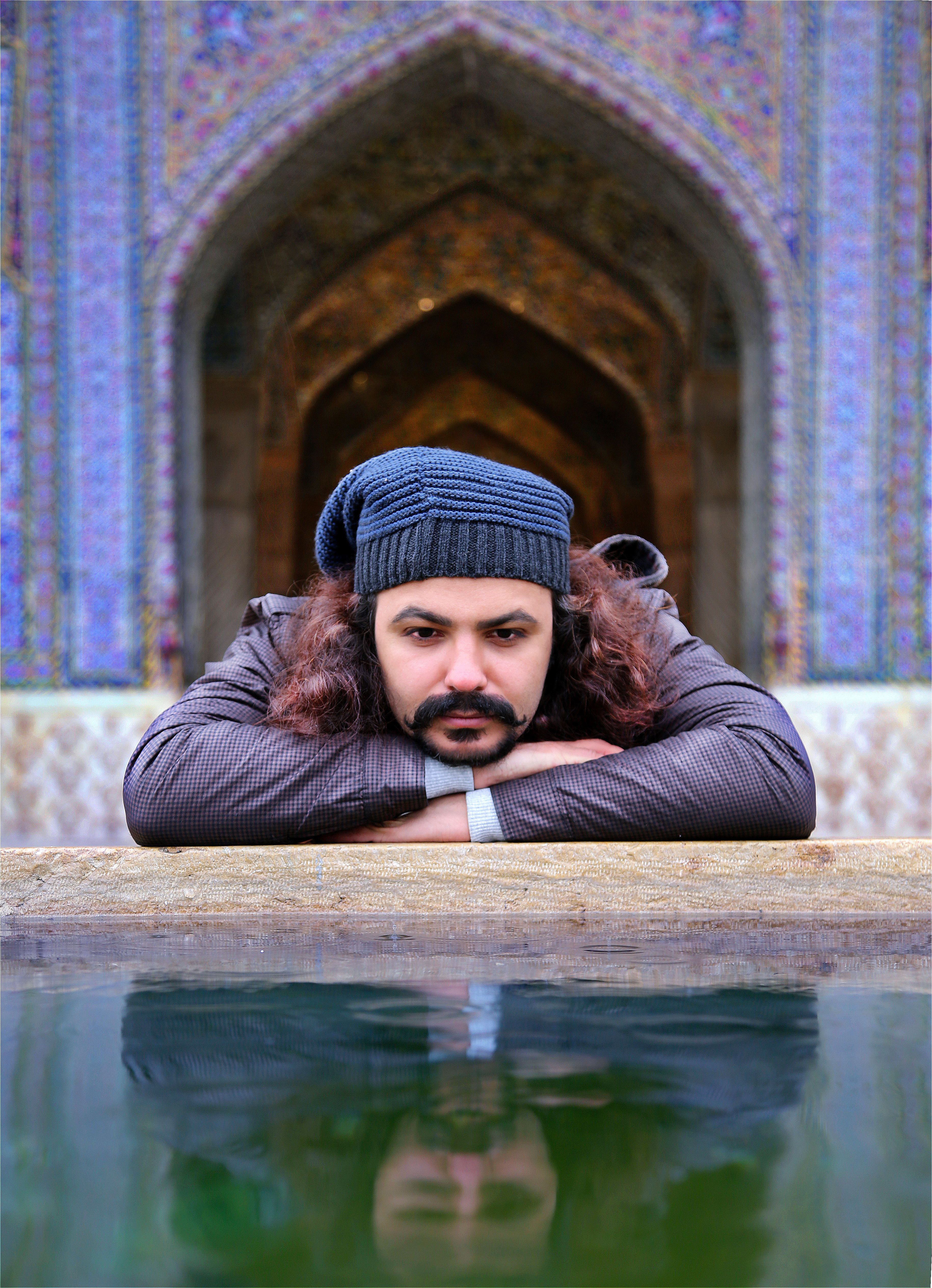A contemplative man in a knit hat gazes into a reflective pool by an ornate archway.