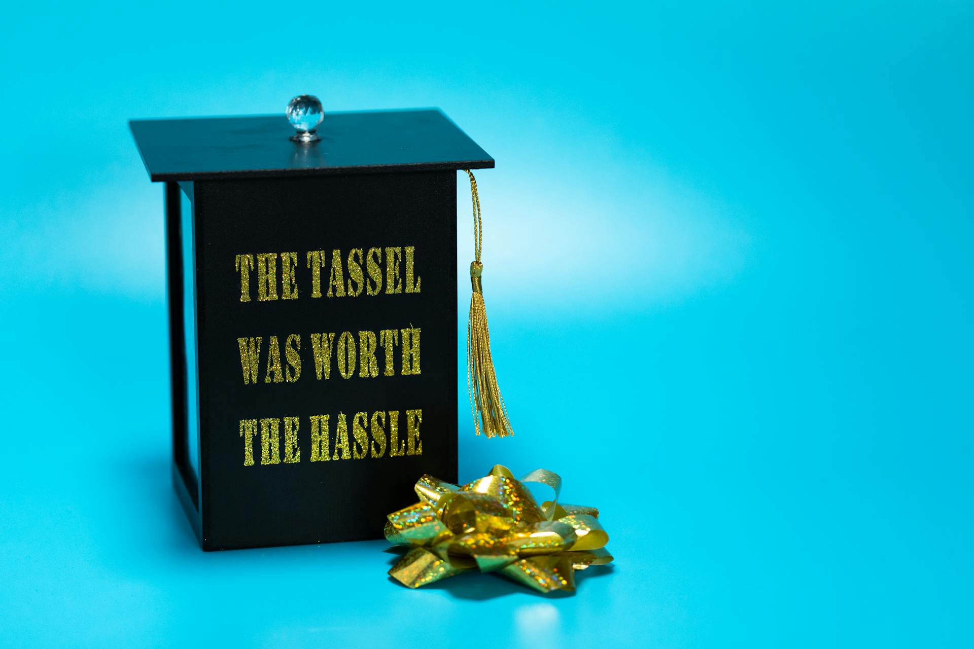 A black graduation cap box with a gold tassel and decorative bow on a light blue background.