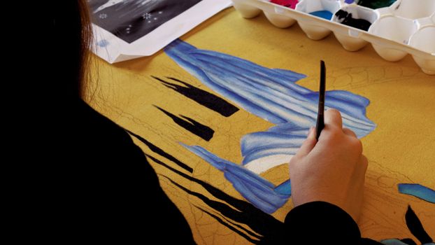 Close-up of an artist painting on a yellow canvas using a detailed blue design.