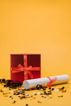 Red gift box and diploma with ribbons on yellow backdrop, celebrating achievement.